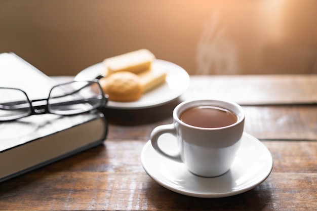 Taza de café caliente con vapor y bocadillos de obleas, viejo libro grueso con vasos en el piso de la mesa vieja y luz de la ventana borrosa en la sala de estar