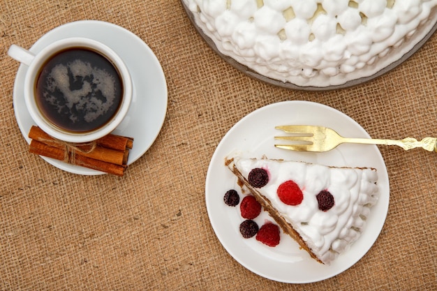 Taza de café, canela, tenedor y rebanada de bizcocho decorado con crema batida y frambuesas en mesa con tela de saco. Vista superior