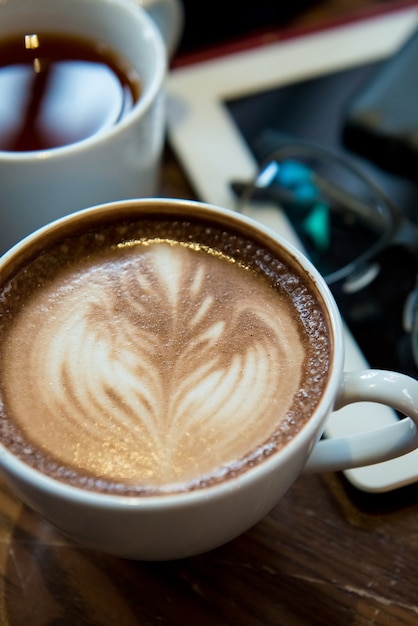 Una taza de café Cappuccino puso en la mesa de madera con tableta, vasos y té en el fondo.