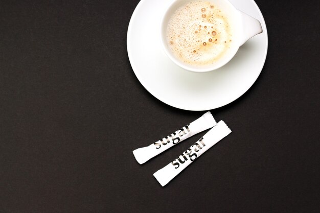 Taza de café capuchino con sobres de azúcar blanco sobre mesa negra.