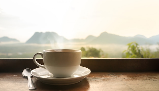 Taza de café de cerámica blanca en mesa de madera en la mañana con la luz del sol sobre el paisaje borroso de las montañas