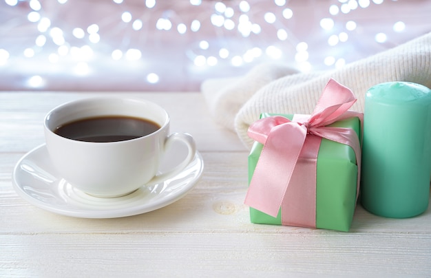 Una taza de café de cerámica blanca, un regalo y una vela sobre un fondo claro con una guirnalda encendida.