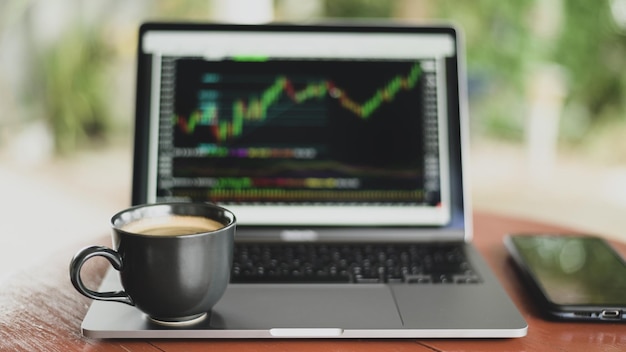 Taza de café colocada en la computadora portátil con gráfico de valores en pantalla, tomada desde el frente.