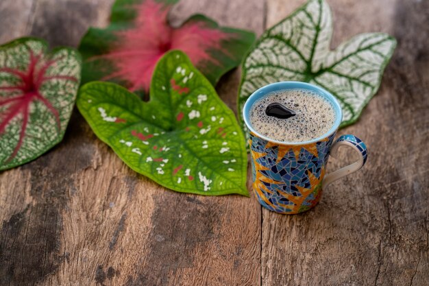 Taza de café contra hojas de Caladium multicolor sobre mesa de madera con textura