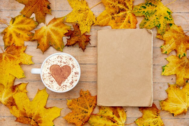 Taza de café con un corazón de canela y libro sobre la mesa, hojas de arce alrededor