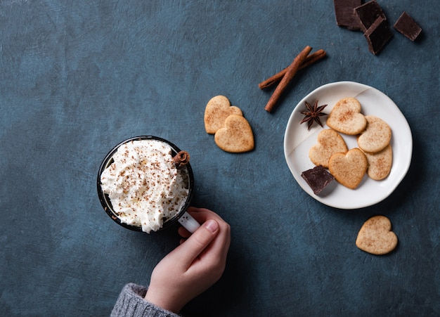 Una taza de café con crema y chispas de chocolate en la mano sobre una mesa azul oscuro con galletas caseras, chocolate y canela. Vista superior y espacio de copia