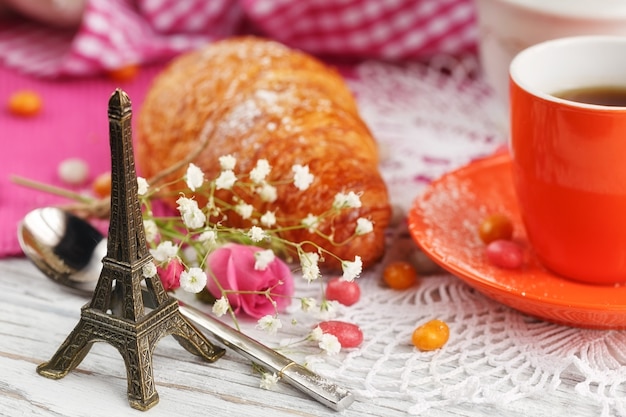 Taza de café y croissant están decorados por la pequeña Torre Eiffel, servilletas, rosas y dulces en una mesa de madera blanca