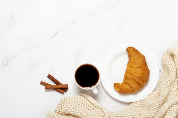 Taza de café, croissant en un plato y una bufanda tejida, palitos de canela sobre una mesa de mármol