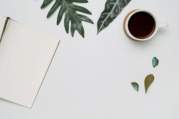 Taza de café y cuaderno sobre fondo blanco de madera con hojas de monstera tropical