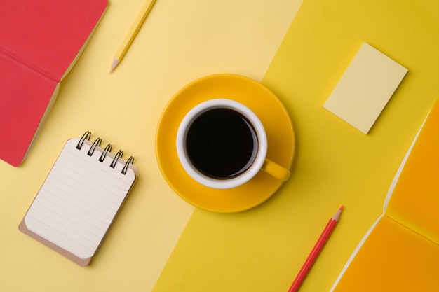 Taza de café, cuadernos y lápices sobre fondo amarillo.