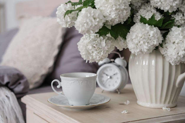 Una taza de café, un despertador y un jarrón de flores blancas en la mesita de noche Hora del té Hora del café