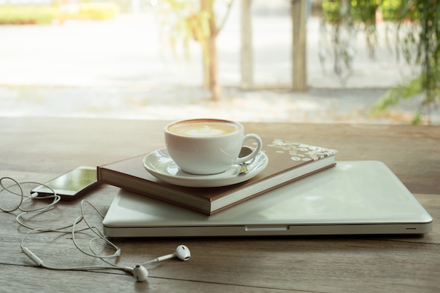 Taza de café encima del libro y del ordenador portátil en la tabla de madera con el teléfono celular.