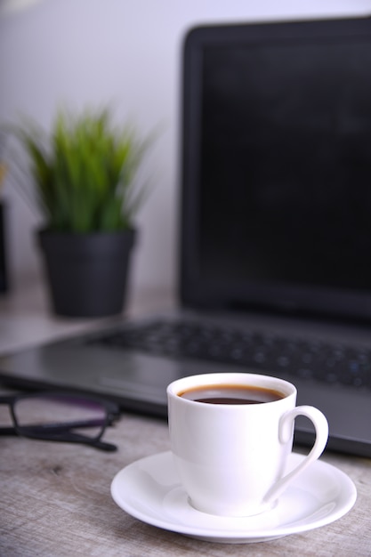 Taza de café en escritorio de oficina con ordenador portátil, computadora en la mesa de madera. De cerca, con espacio para tu texto