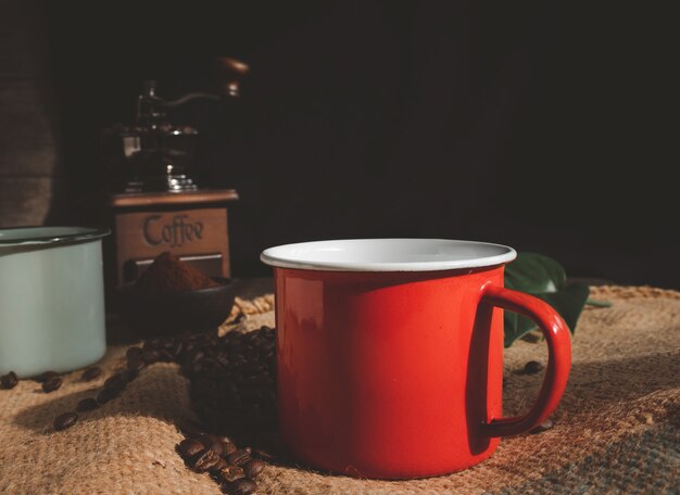 Taza de café de esmalte rojo, café molido, granos de café tostados, molinillo y hojas de monstera en la mesa de madera con fondo de arpillera