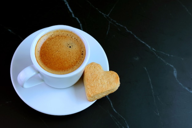 Taza de café expreso con una galleta de mantequilla en forma de corazón sobre mesa negra