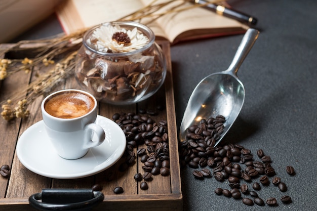 La taza del café express, el grano de café, el libro y las flores secas sacuden en la bandeja de madera con mañana caliente.