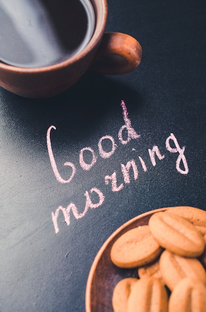 Taza de café y de galleta en un fondo oscuro. Inscripción buenos días.