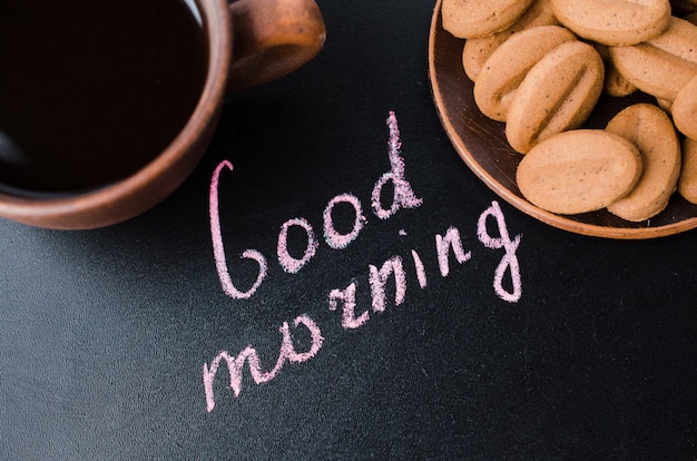 Taza de café y de galleta en un fondo oscuro. Inscripción buenos días.