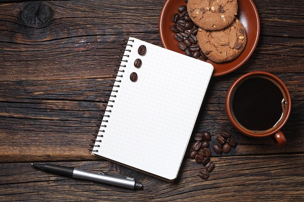 Taza de café, galletas y bloc de notas abierto sobre una mesa de madera, vista superior. Espacio para tu texto