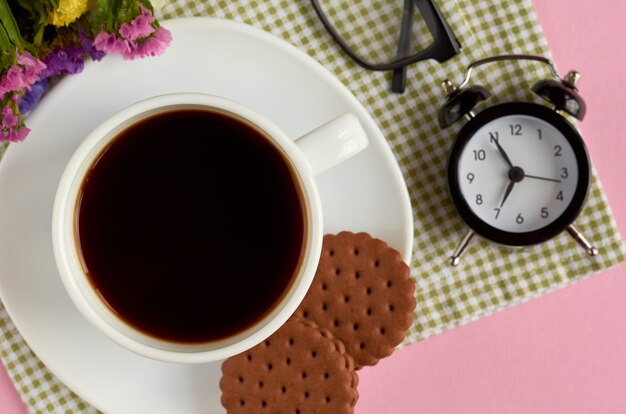 Taza de café con galletas, reloj despertador, flores, rímel, sobre superficie rosa.