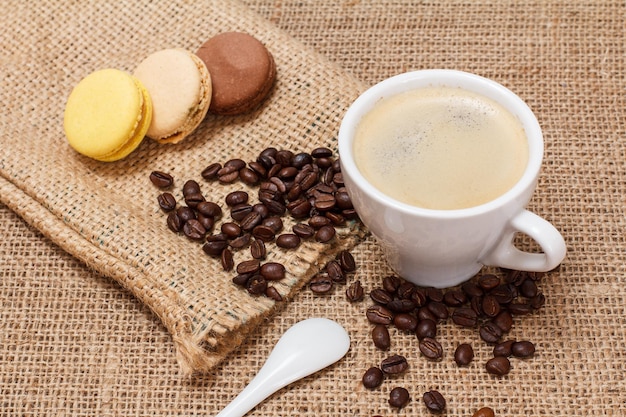 Taza de café, granos de café con saco de lona y macarrones sobre fondo de tela de saco. Vista superior.