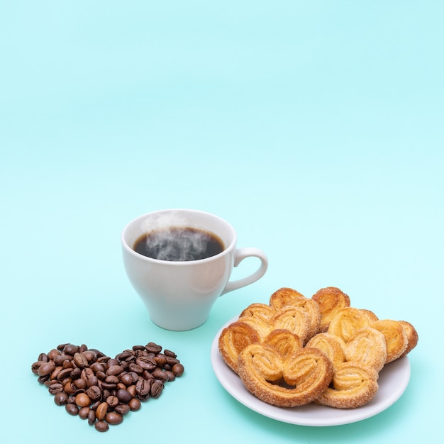 Taza de café humeante, galletas en forma de corazón, granos de café en forma de corazón sobre un fondo azul.