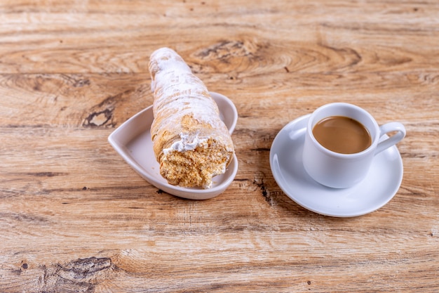 Una taza de café junto a un delicioso hojaldre aromático con crema proteica, un delicioso desayuno o merienda en mesa de madera.