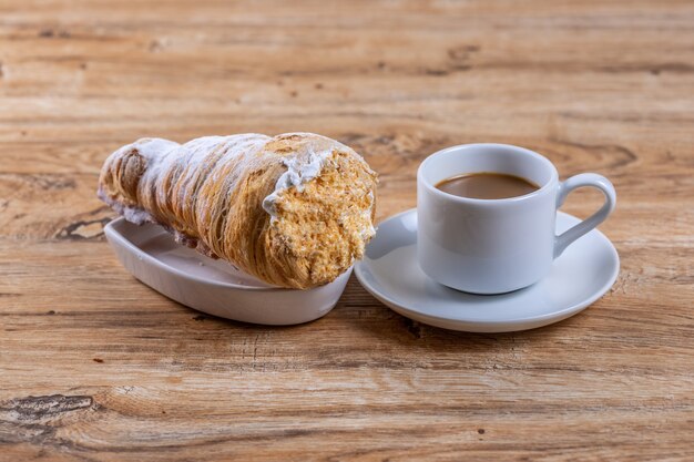 Una taza de café junto a un delicioso hojaldre aromático con crema proteica, un delicioso desayuno o merienda en mesa de madera.