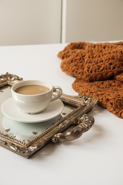 Taza de café con leche en bandeja dorada vintage y manta tejida a cuadros marrón sobre fondo blanco.