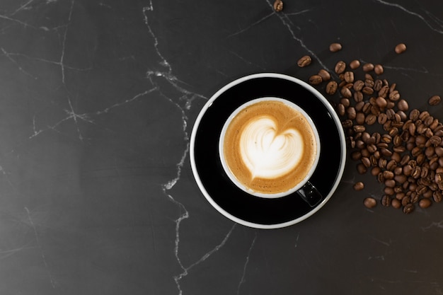 Taza de café con leche caliente con hermosa textura de arte latte de espuma de leche con grano de café sobre fondo de mármol negro. Vista aérea, copie el espacio. Publicidad para menú de cafetería. Menú de cafetería.