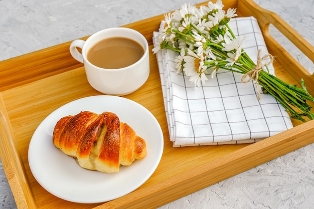 Taza de café con leche, croissant recién horneado, servilleta a cuadros y flores de manzanilla en bandeja de madera