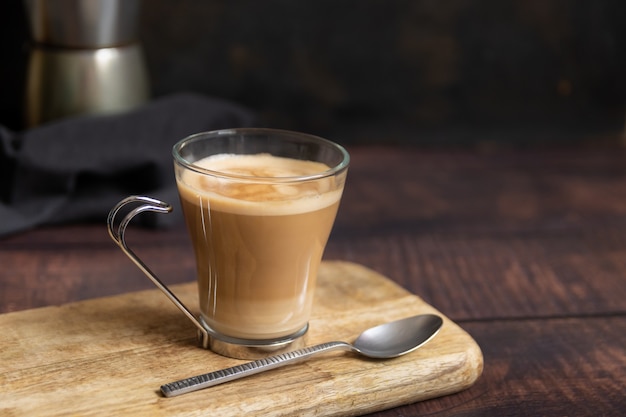 Taza de café con leche y cucharadita de mesa de madera y cafetera italiana en el fondo