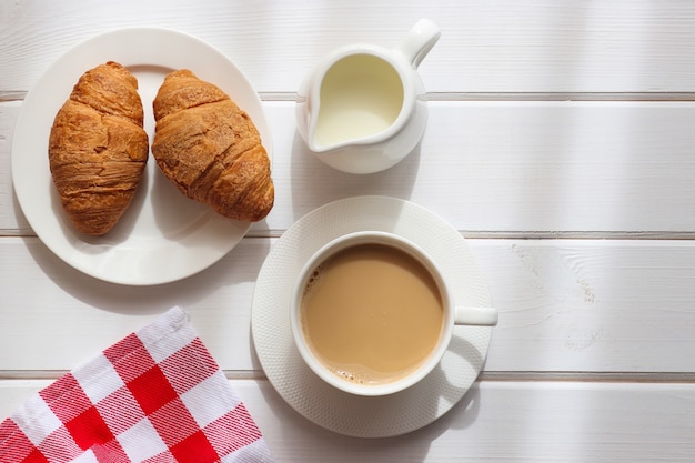 Taza de café con leche y dos croissants en un plato sobre la mesa de madera blanca con sombra de luz