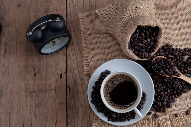 Taza del café con leche con los granos de café y el despertador en fondo de madera