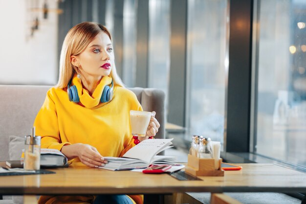 Taza con café con leche. Hermosa mujer de ojos azules sosteniendo una taza con sabroso café con leche mientras está sentado en la cafetería