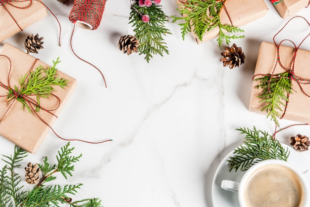 Taza de café con leche con regalo de Navidad o caja de regalo envuelta en papel kraft, decorada con ramas de árboles de navidad, conos de pino, bayas rojas, en mesa de mármol blanco, marco de espacio de copia