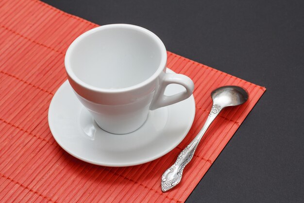 Taza de café con leche vacía, platillo y cuchara sobre una servilleta de bambú rojo con fondo negro. Vista superior.
