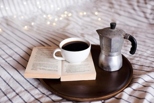 Taza de café en libro abierto con cafetera en la cama de cerca. Buenos días.