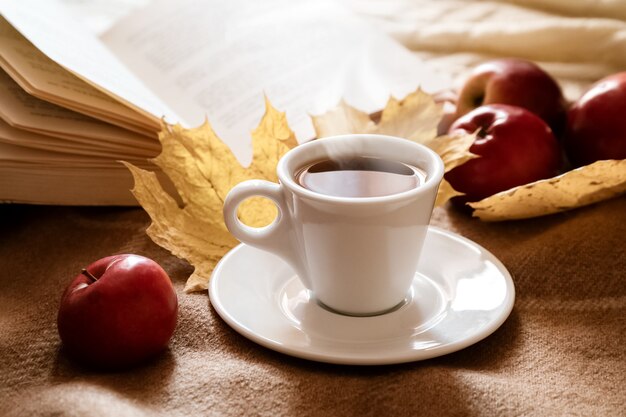 Taza de café llena de espresso caliente sobre una manta de lana beige cerca de un libro abierto y manzanas rojas dispersas