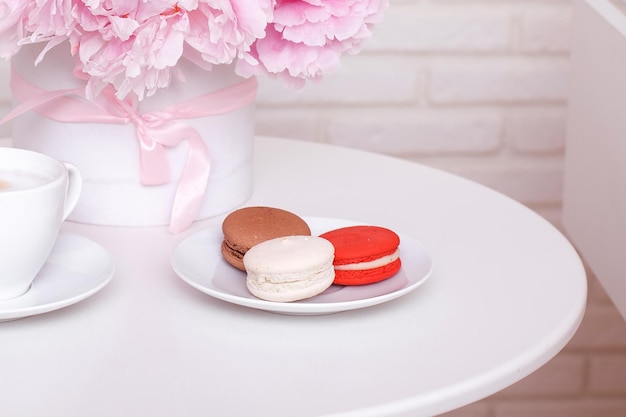 Taza de café, macarrones dulces y peonías rosas sobre fondo blanco de mesa