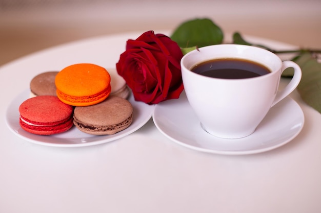 Taza de café, macarrones dulces y rosa roja sobre fondo blanco de mesa