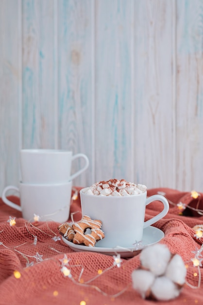 Taza de café con malvavisco y galletas de estrellas sobre una cálida manta de coral tejida, luces navideñas y flores de algodón.