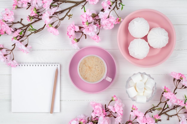 Taza de café con malvavisco por la mañana para el desayuno. Cuaderno en blanco con bolígrafo. Diario, concepto de planificación. Maqueta de primavera romántica con espacio de copia. Flor de cerezo sobre fondo blanco de madera. Endecha plana.