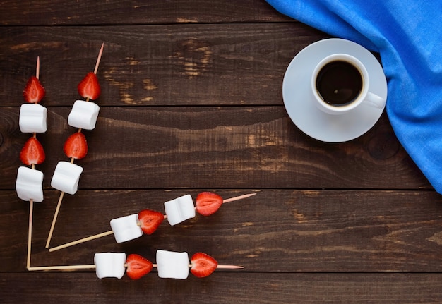 Una taza de café y malvaviscos con fresas frescas en brochetas sobre una mesa de madera. La vista superior