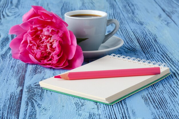Taza de café de la mañana, cuaderno vacío, lápiz y flores de peonía blanca en la mesa de madera azul