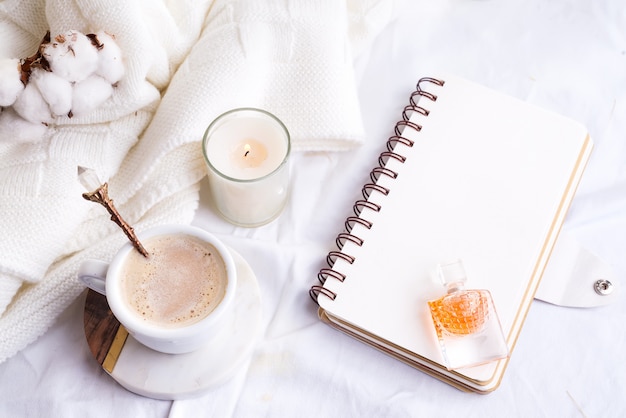 Taza de café de la mañana con flores de cuaderno, vela y algodón en una vista superior de cama blanca