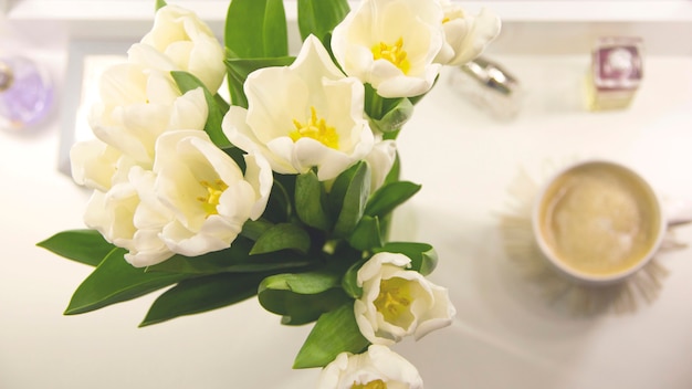 Una taza de café en la mañana junto a un ramo de tulipanes blancos.