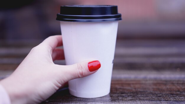 Taza de café en una mesa de madera en manos femeninas. Vaso de papel Tapa de plástico. Hermosas manos con uñas rojas