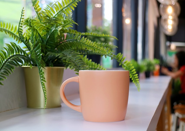 Taza de café naranja pastel con helechos en macetas en una mesa larga con gente borrosa sentada en la distancia