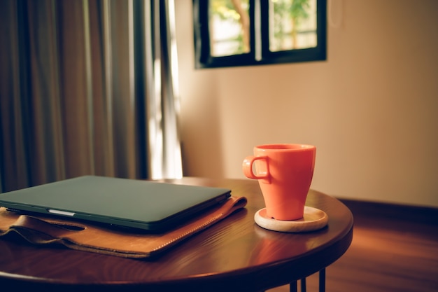 Taza de café naranja y portátil sobre la mesa de la cafetería.